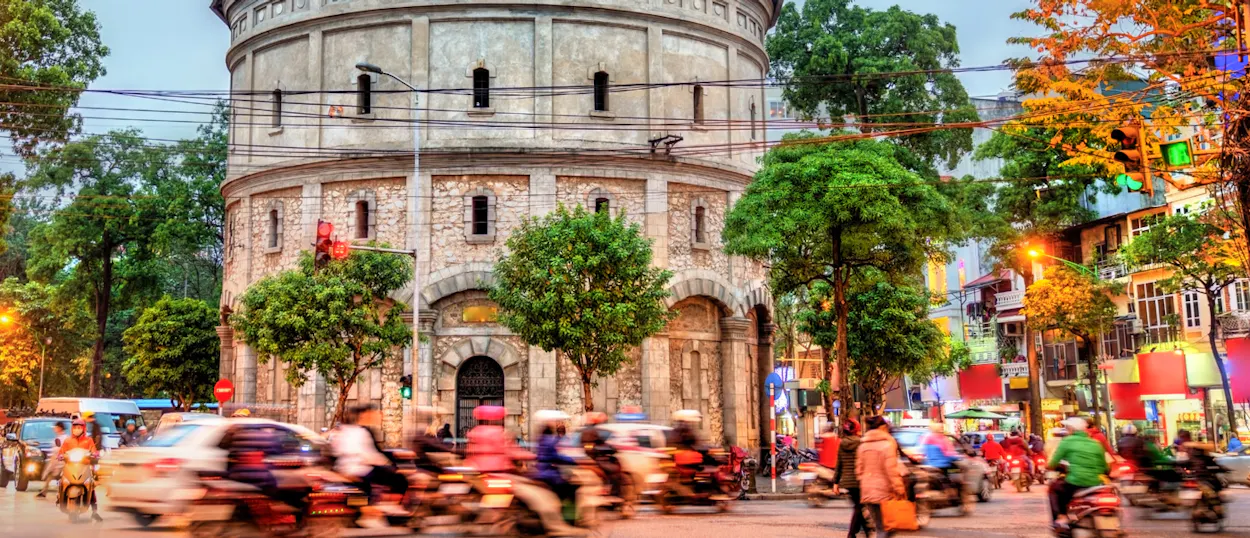 Bustling Hanoi street at dusk, with a historic water tower and blurred motorbikes speeding past.