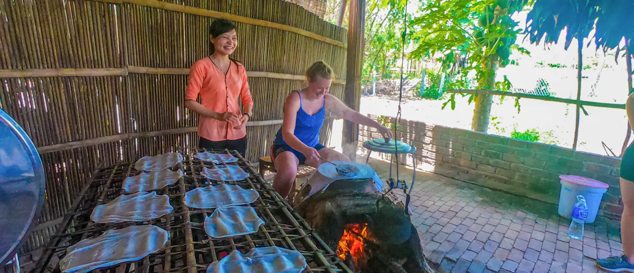 A smiling local guide teaches a tourist to cook traditional food over an outdoor fire in Hoi An.
