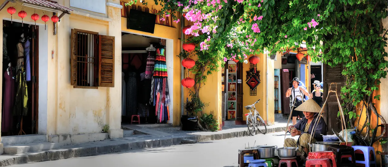 A woman in a conical hat sells food on a charming street in Hoi An, Vietnam, with colorful shops.