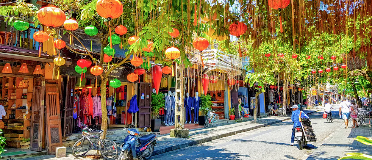 A charming street in Hoi An, Vietnam, adorned with colorful lanterns, green trees, and people walking.