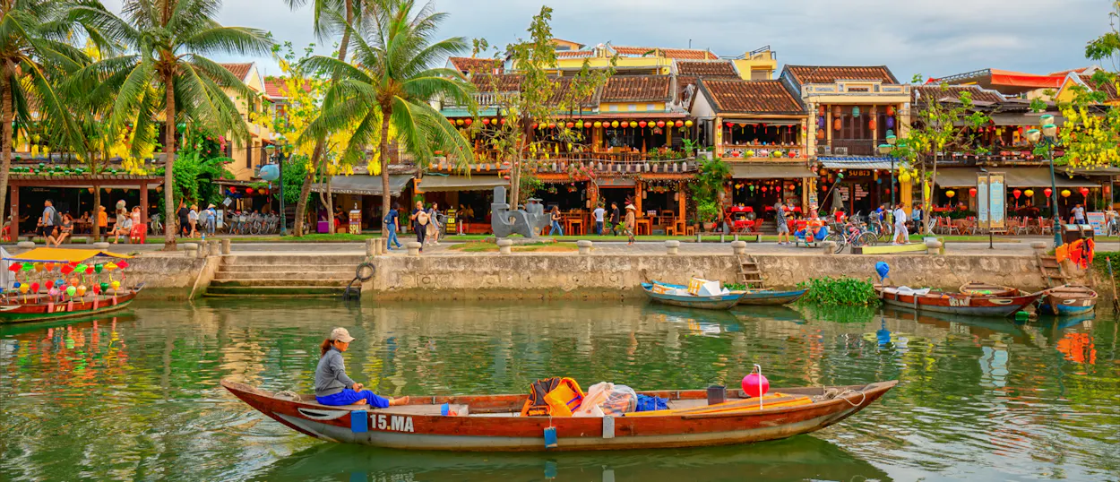 A person rows a boat on a river with colorful buildings and palm trees in Hoi An.